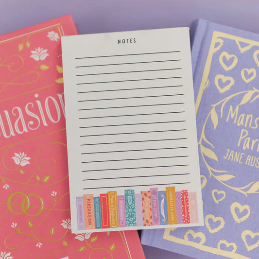 Notepad with striped lines and on the bottom various books of different sizes and colors are lined up all of which are written by Jane Austen. 