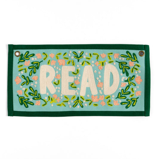 A rectangular, light blue canvas banner with the word "READ" embroidered in off-white, surrounded by embroidered pink flowers and green leaves, with a dark green border and gold grommets at the top corners.
